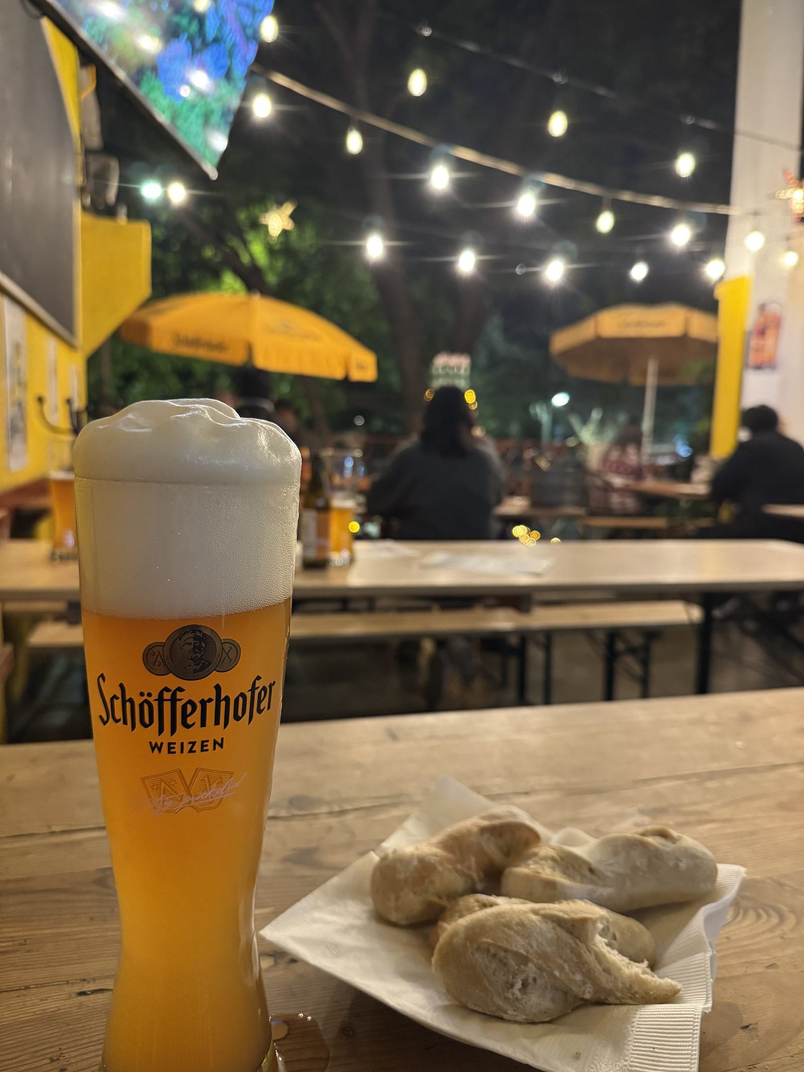 A glass of wheat beer on a wooden table at an outdoor beer garden at night, string lights above