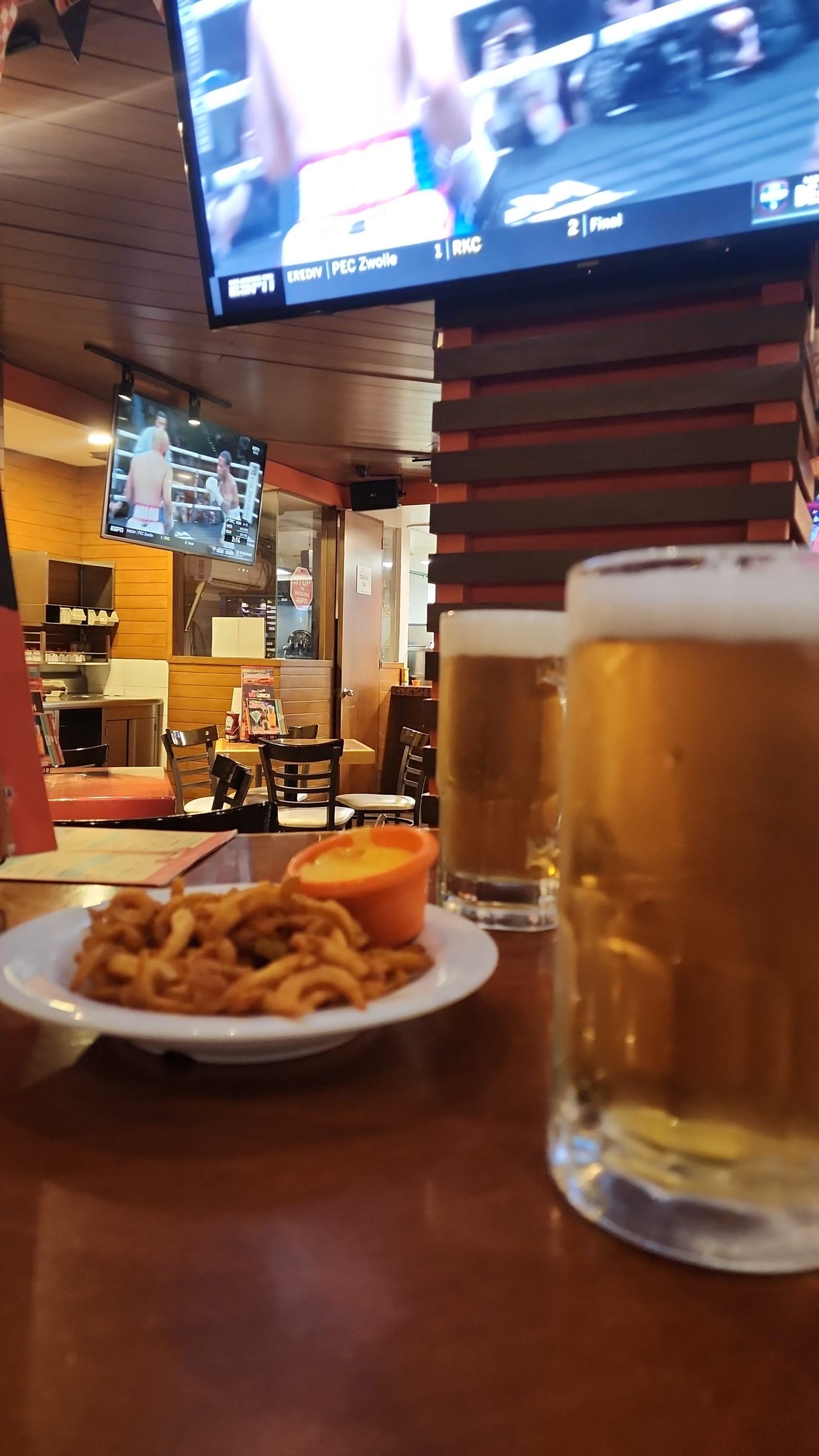A sports bar table with a football match on a television screen, beer mugs and a plate of curly fries